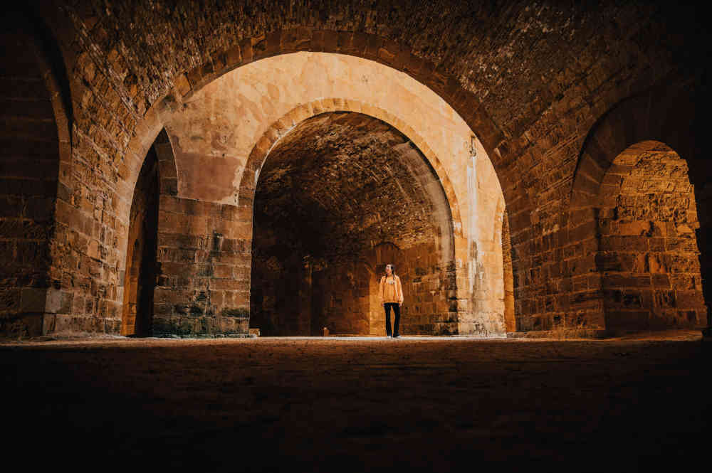 Large arched brick catacomb-like caverns/tunnels, orange illuminated, and with a central standing woman looking at her surroundings.